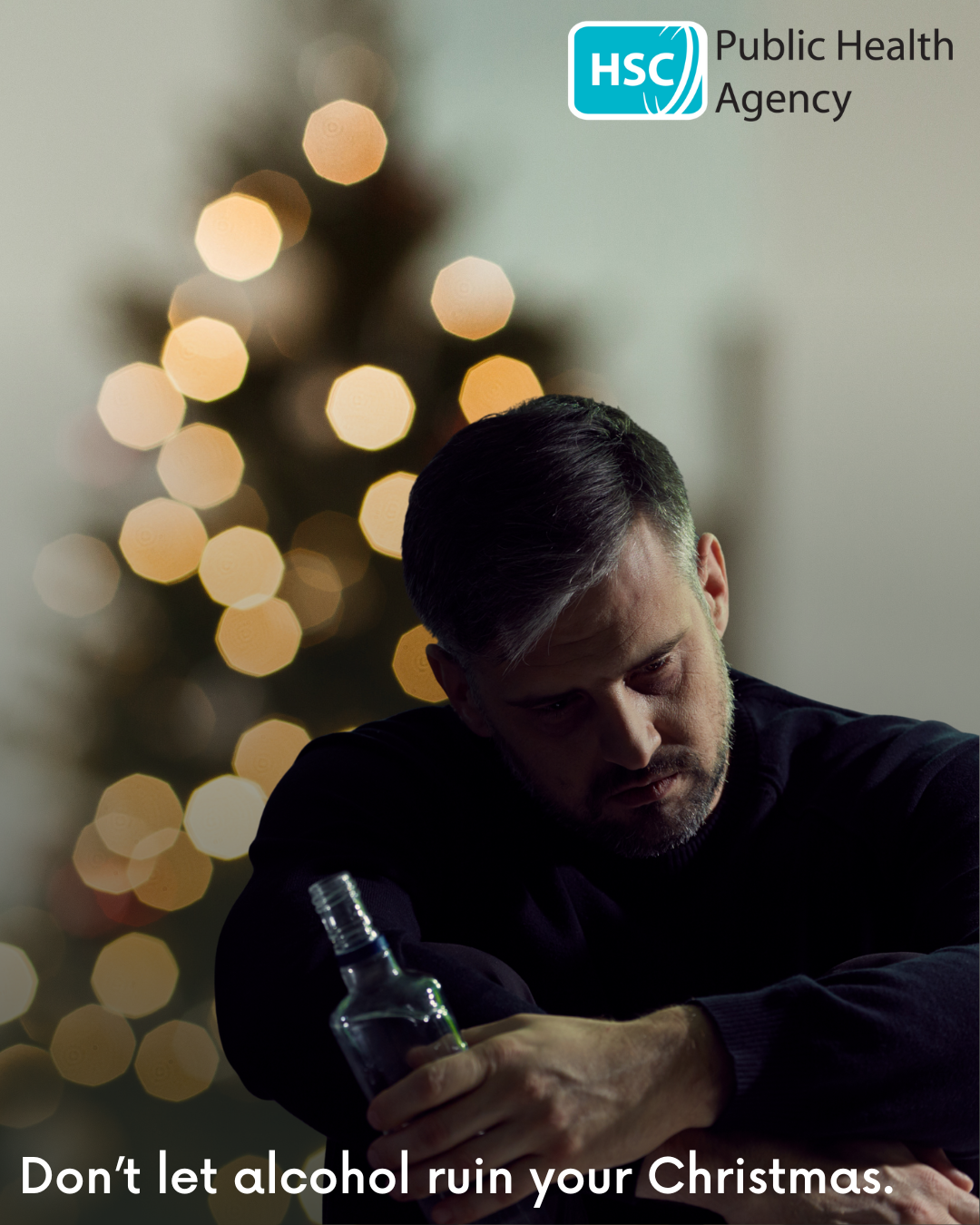 Photograph of a man holding a bottle of alcohol in front of Christmas tree, with a sad expression on his face.