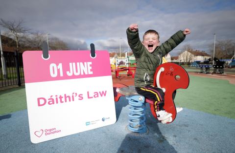 Dáithí Mac Gabhann on a red rocking horse his hands in the air with: "01 June, Dáithí's Law" written on a placard beside him  