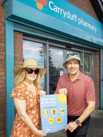 Photograph of Frances Dowds, Health Improvement Manager at PHA with Garth Newberry, Community Pharmacist, holding the Care in the Sun poster outside Carryduff Pharmacy. 
