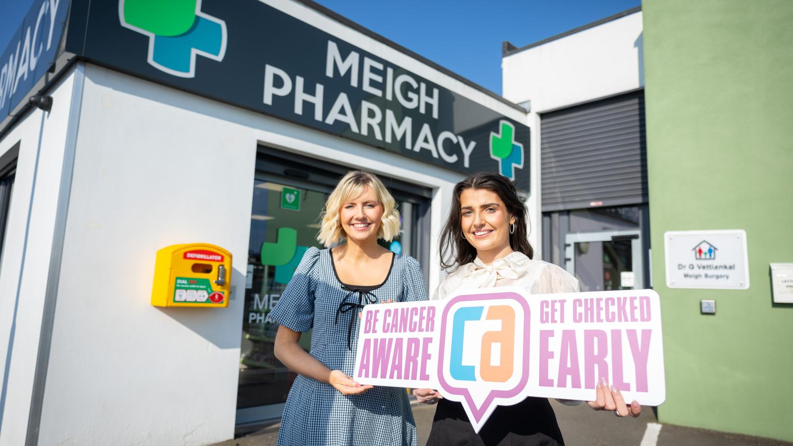 Photograph of Dr Anna Cullen and Clare Conroy (pharmacist) standing outside Meigh pharmacy, holding a 'Be Cancer Aware' sign