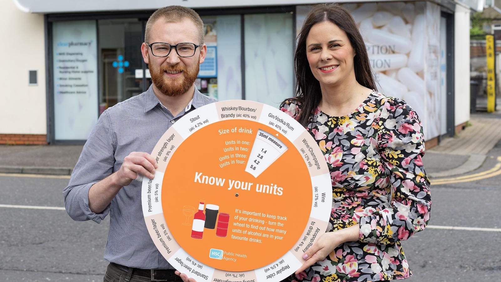 Photograph of PHA rep and pharmacist, holding the 'know your units' campaign unit calculator wheel, outside Clear Pharmacy in Bangor. 