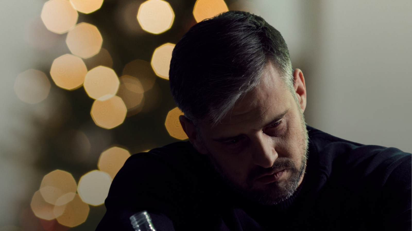 Photograph of a man holding a bottle of alcohol in front of Christmas tree, with a sad expression on his face.
