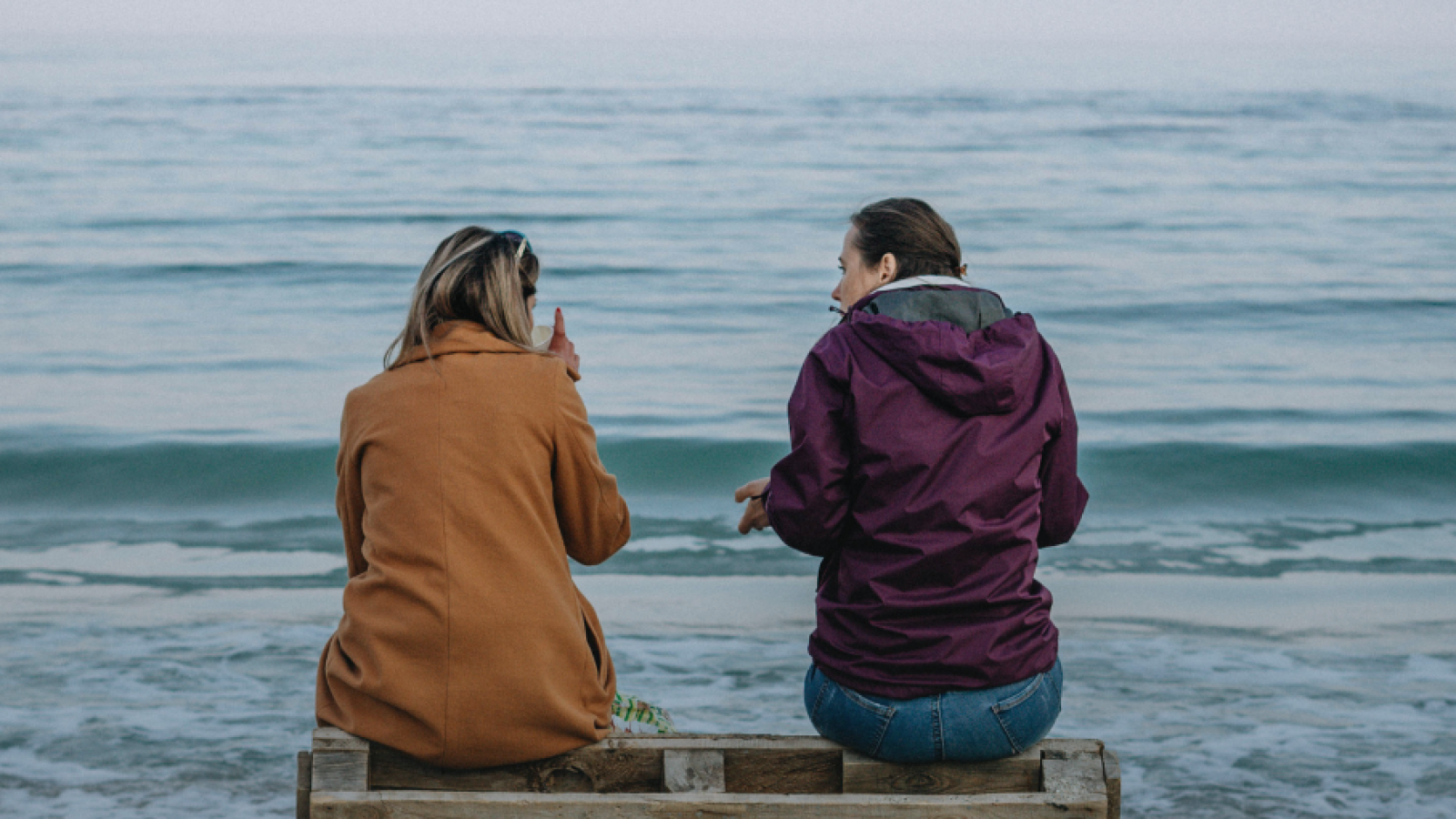 Photograph of two people sitting by the beach talking. With text stating "1 in 2 of us will develop some form of cancer in our lifetime." To highlight World Cancer Day 2026.