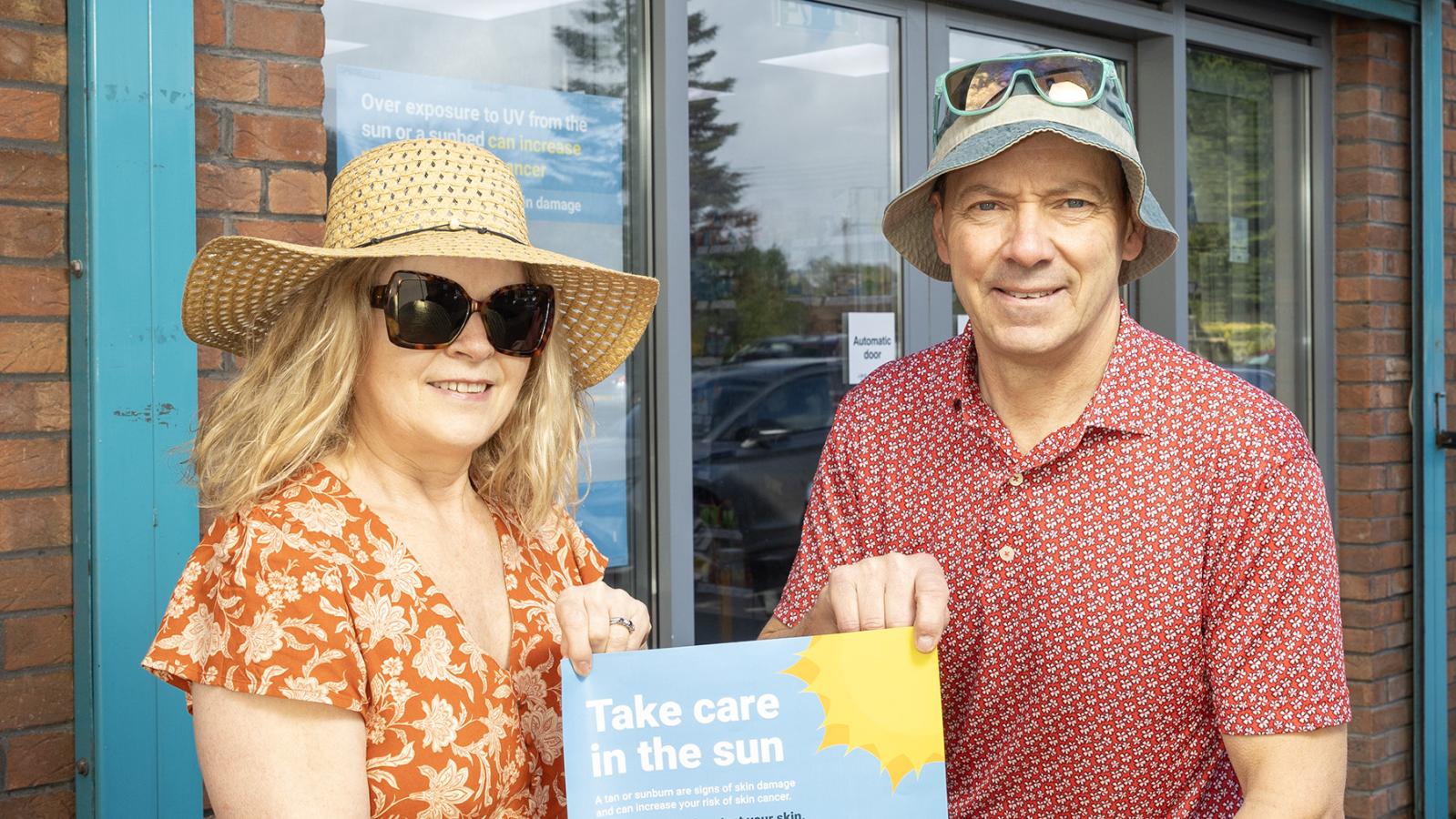 Photograph of Frances Dowds, Health Improvement Manager at PHA with Garth Newberry, Community Pharmacist, holding the Care in the Sun poster outside Carryduff Pharmacy. 